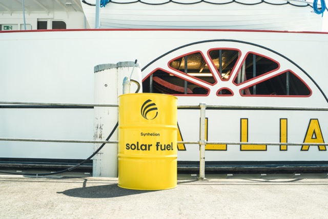 Yellow Synhelion renewable diesel barrel positioned in front of a historic steamboat, with visible deck details and classic vessel features in the background