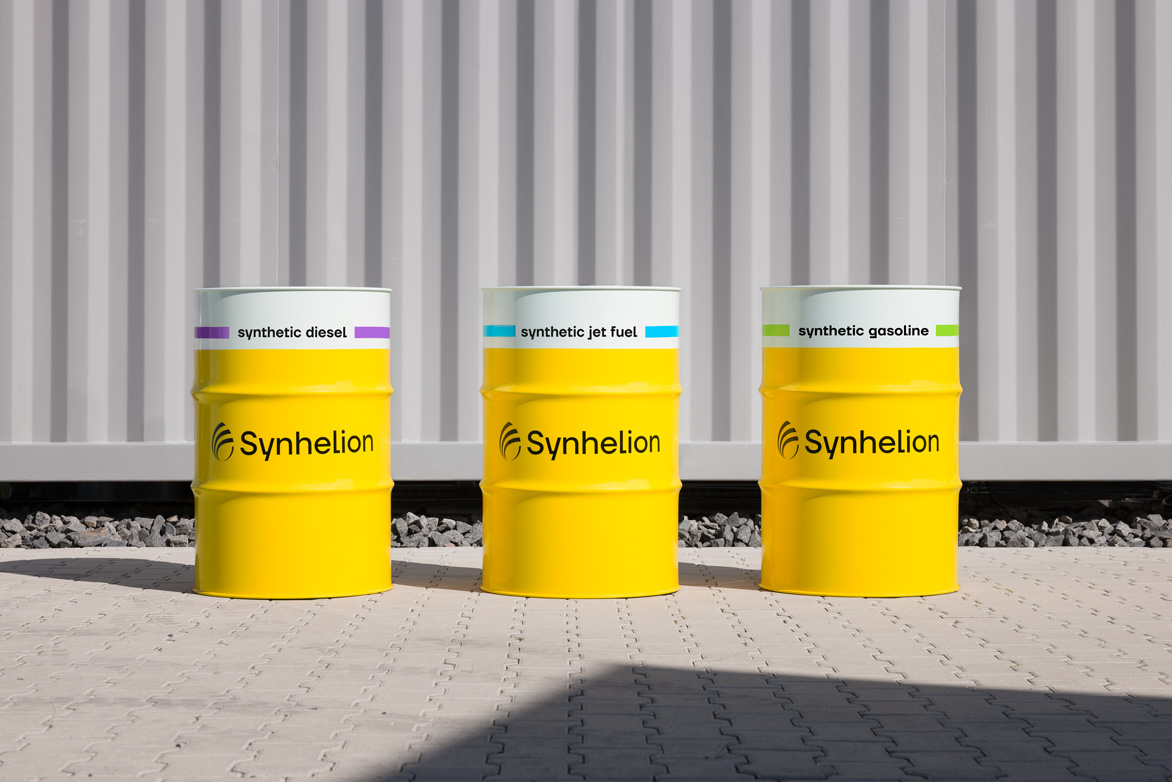Three yellow fuel barrels labeled “Synhelion”, placed outdoors on a paved surface in front of a corrugated metal wall.
