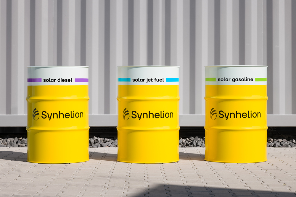 Three yellow fuel barrels labeled &ldquo;Synhelion&rdquo;, placed outdoors on a paved surface in front of a corrugated metal wall.