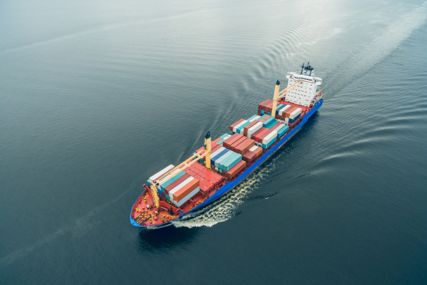 Aerial view of a cargo container ship sailing on calm open water, carrying stacked multicolored containers with a visible wake behind it.