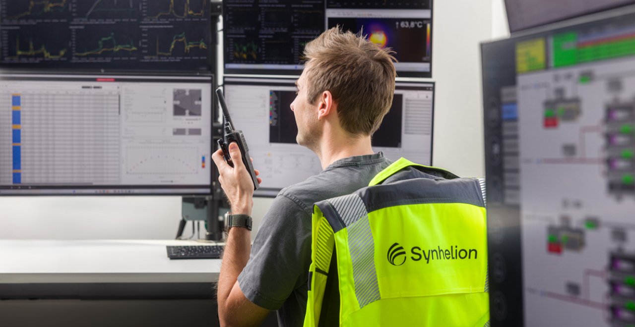 Technician wearing a high-visibility Synhelion vest monitoring systems on multiple control screens, using a handheld device in a control room.