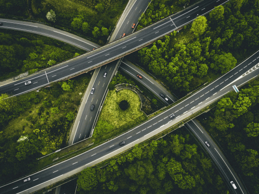 Aerial view of a highway interchange with multiple overpasses crossing through dense green forest, showing curved roads and light traffic.